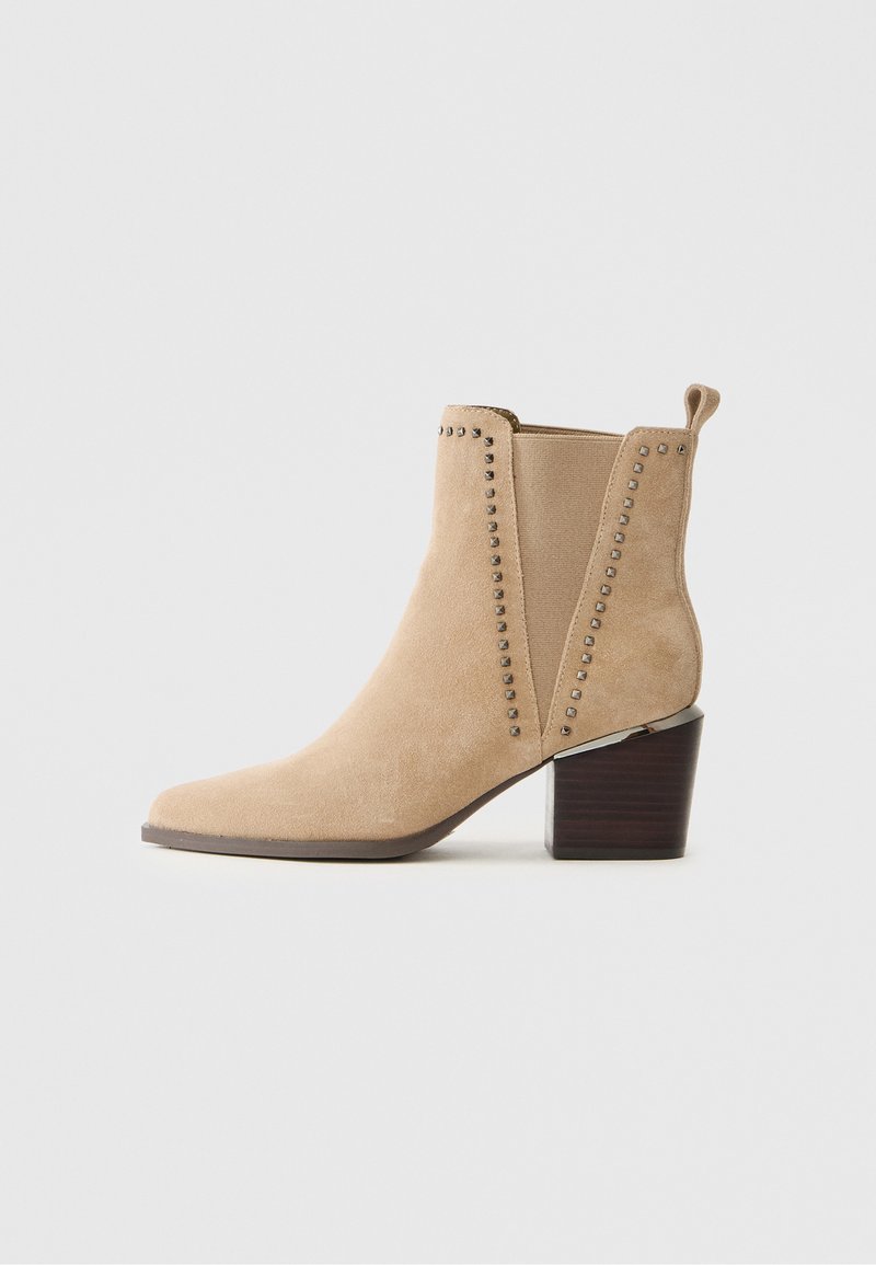Beżowe zamszowe botki za kostkę z szpiczastym noskiem, ozdobione ćwiekami po bokach, na solidnym drewnianym obcasie. Posiadają pętelkę do łatwego zakładania.