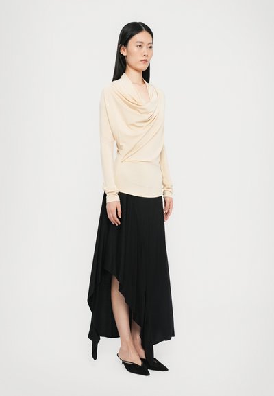 Beige gedrapeerde top met lange mouwen gecombineerd met een zwarte asymmetrische rok, met zachte stoftexturen en puntige zwarte mules.