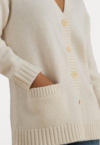 Cardigã creme feito de tecido de malha texturizado, com decote em V, bordas caneladas, quatro botões claros e dois bolsos frontais.