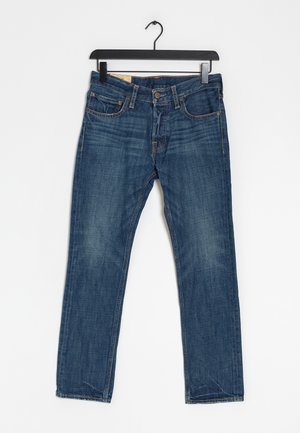 Jean en denim bleu avec poches avant et fermeture à boutons, suspendu à un cintre noir sur un fond blanc.