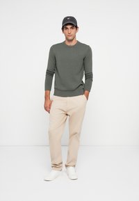 Mann med olivengrønn genser, beige bukser, hvite sneakers og mørk grå caps som står mot hvit bakgrunn.