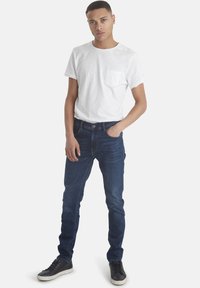 Vit t-shirt med rund hals och bröstficka, kombinerad med slim-fit mörkblå jeans och svarta sneakers med vit sula.
