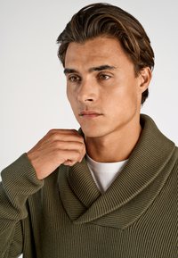 Olijfgroene ribbelgebreide trui met een sjalkraag, gedragen over een wit t-shirt. De hand van het model houdt de kraag vast.