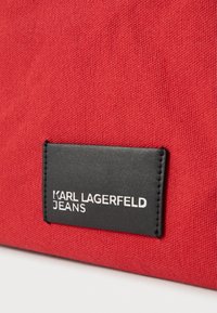 Czerwona torba materiałowa z czarną prostokątną etykietą z wytłoczonym napisem "KARL LAGERFELD JEANS". Teksturowana powierzchnia, kształt prostokątny.