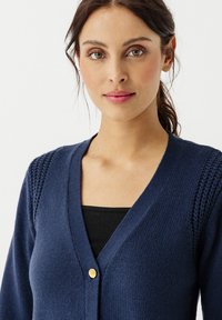 Cardigan lavorato a maglia blu navy con scollatura a V, dotato di dettagli testurizzati sulle spalle e chiusura con un singolo bottone dorato. Design semplice e aderente.