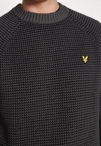 Knitted black sweater with a textured pattern, featuring a ribbed round neckline and a small yellow eagle logo on the chest.