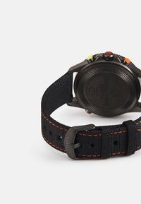 Bracelet de montre en tissu noir avec des coutures orange, boucle en acier inoxydable et boîtier noir texturé avec des boutons colorés et des détails gravés.