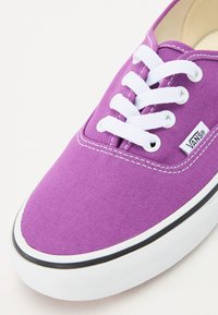 Purple canvas sneaker featuring white laces, black trim along the sole, and a small branded tag on the side. Textured surface for grip.