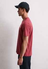 Rode katoenen t-shirt met korte mouwen, ronde halslijn en gouden logo. Ggedragen met marineblauwe broek en een zwarte pet, vanuit de zijkant bekeken.