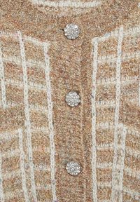 Tissu tricoté texturé beige et blanc avec une bande verticale comprenant trois boutons argentés en forme de fleur incrustés de strass clairs.