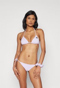 Lilla trekant bikini med tynne stropper og en liten logo på toppen. Buksene har knyting på siden med en matchende lilla scrunchie på håndleddet.