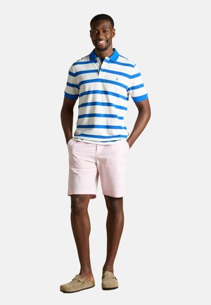 Smilende mand stående med hænderne i lommerne iført en hvid og blåstribet polo skjorte, lyserøde shorts og beige slip-on sko.