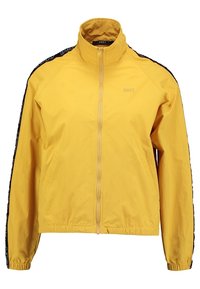 Żółta kurtka przeciwdeszczowa z wysokim kołnierzem, pełnym zamkiem błyskawicznym i czarnymi paskami bocznymi z napisem. Materiał wydaje się lekki i gładki.