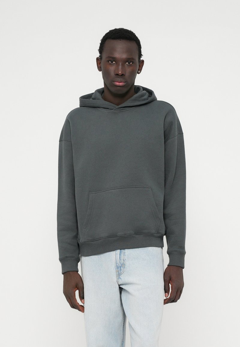 Pelēka kapucēta džemperis ar priekšējo kabatu, brīva piegriezuma, ar ribotām aprocēm un apakšmalu, savienots ar gaiši ziliem džinsiem.