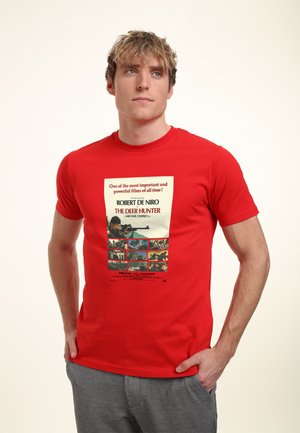 Rød bomulds t-shirt med et trykt filmmuseum af "The Deer Hunter," med en hvid baggrund, sort tekst og farverige billeder arrangeret nedenfor.