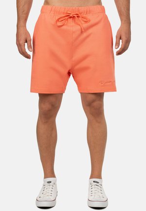 Mężczyzna w koralowych szortach ze sznurkiem i białych niskich trampkach, stojący na jednolitym tle.