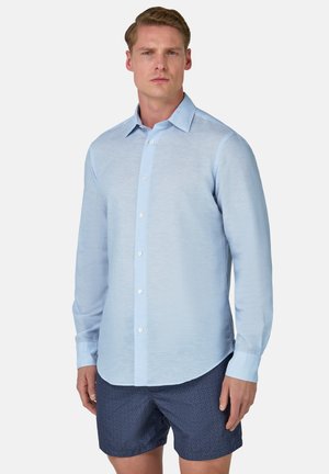 Camicia a maniche lunghe azzurra chiara con chiusura a bottoni, colletto aperto e tessuto leggermente texturizzato. Abbinata a pantaloni corti navy a fantasia.