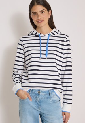 Donna che indossa una felpa bianca con strisce orizzontali nere e cordini blu, abbinata a jeans azzurri, in piedi davanti a uno sfondo semplice.