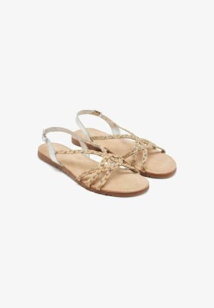 Sandalias planas beige con tiras trenzadas, detalles en plata y una suela acolchada. Presenta un diseño minimalista y correas ajustables en los tobillos.