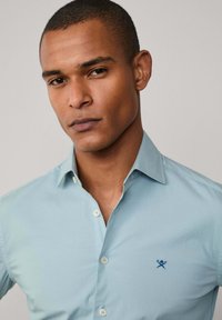 Lichtblauwe button-up shirt van gladde katoen. Beschikt over een klassieke kraag en een geborduurd marineblauw logo op de linkerborst.