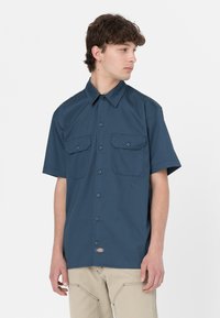 Camicia a maniche corte con bottoni in tessuto blu scuro, dotata di due tasche sul petto, colletto a punta e un piccolo cartellino con logo sull'orlo.