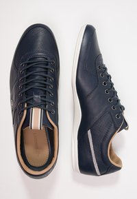 Par de zapatillas de cuero azul marino con suelas blancas, forro interior beige y una lengüeta de tela a rayas en el talón, mostradas desde vistas superior y lateral.