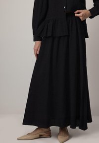 Žena v černé dlouhé sukni a odpovídajícím černém topu s volánovým detailem, béžových špičatých slingback balerínách, stojící na jednobarevném pozadí.