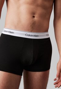 Schwarze Baumwoll-Boxershorts mit einem weißen elastischen Bund, der das "Calvin Klein"-Logo trägt. Glatte Textur und enganliegendes Design.
