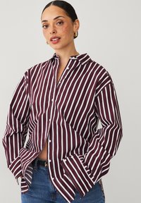 Maroon and white striped button-up shirt with a collar, relaxed fit, and long sleeves. Features a front pocket and split at the hem.