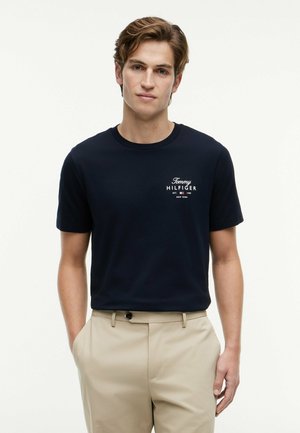 Ung mand med lysebrunt hår iført en sort Tommy Hilfiger t-shirt og beige bukser, stående med hænderne i lommerne mod hvid baggrund.