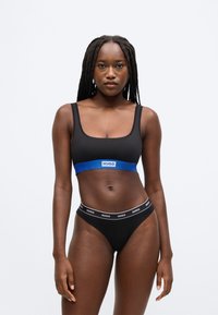 Musta rintaliivit sinisellä HUGO-logonauhalla, yhdistettynä mustaan thong-alushousuun, jossa valkoinen logovyötärönauha. Sileä kangas ja yksinkertainen muotoilu.