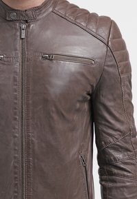 Giacca in pelle marrone con dettaglio trapuntato sulle spalle, tasche a zip sul petto e sui lati, e chiusura a zip frontale.