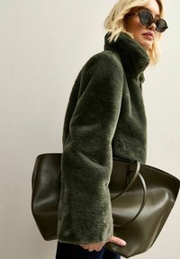 Mujer rubia con chaqueta peluda verde oscuro y gafas de sol sostiene un gran bolso tote de cuero en tono oliva oscuro frente a una pared lisa.