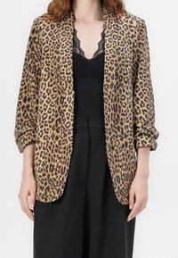 Blazer s leopardím potiskem s hladkou texturou, otevřeným předním designem a ohrnutými rukávy, nošený přes černý krajkový top a černé kalhoty.