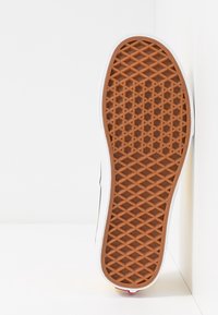 Hnědá gumová podrážka s diamantovým vzorem. Má bílý okraj a červený akcent na patě. Viditelná zpevněná textura pro lepší úchop.