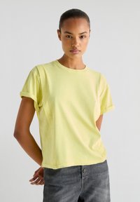 T-shirt a maniche corte, collo rotondo, in tessuto giallo chiaro, con risvolti alle maniche, una vestibilità comoda e una texture morbida. Abbinato a pantaloni grigi.