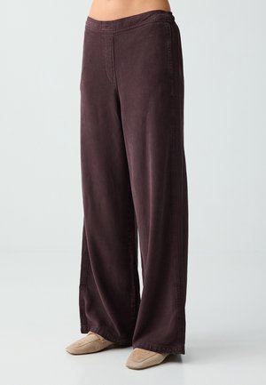 Femme portant un pantalon marron foncé taille haute à jambes larges avec des chaussures plates beiges, debout devant un fond clair uni.