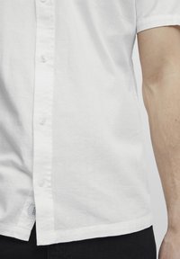 Weißes kurzärmliges Hemd aus leichtem, strukturiertem Stoff. Verfügt über fünf Knöpfe vorne und ein seitliches Etikett.