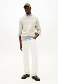 Mand iført beige sweatshirt med emblem, lyseblå skjorte, hvide bukser, sorte sko og olivengrøn kasket, som står med den ene hånd i lommen.