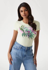 T-shirt ajusté couleur jaune clair avec un motif graphique tropical vert et rose et un design de texte ; associé à un jean en denim bleu taille haute.