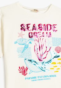 T-shirt couleur crème avec un design vibrant en sequins représentant la vie marine, des coraux, et le texte "RÊVE EN BORD DE MER" en rose et bleu.