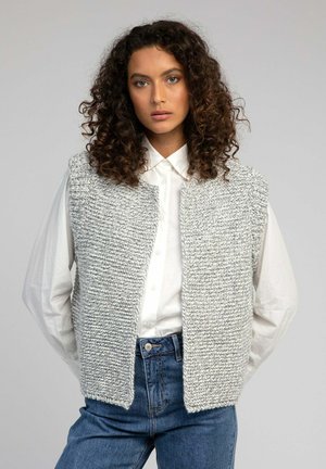Femme aux cheveux bouclés portant un gilet sans manches en tricot gris, une chemise blanche boutonnée et un jean bleu, se tenant devant un fond uni.