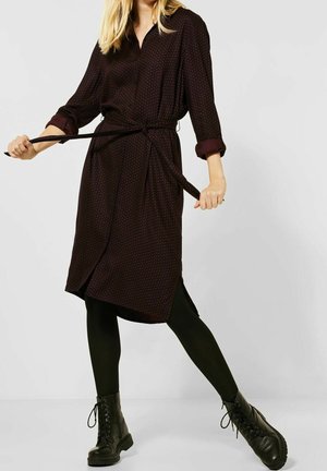 Femme portant une robe foncée à motifs longueur genou avec manches retroussées, des leggings noirs et des bottes noires lacées, tenant la ceinture de la robe tendue.