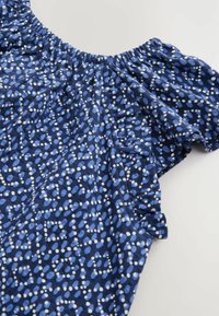 Haut en tissu à motifs bleus avec un col élastique à volants, présentant un mélange de taches bleues claires et foncées ainsi que des points blancs tout au long.