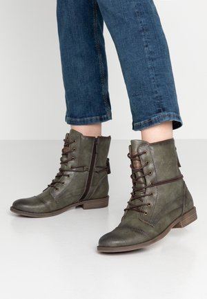 Osoba nosząca oliwkowozielone skórzane botki za kostkę z brązowymi sznurowadłami i zamkami błyskawicznymi, w parze z podwiniętymi niebieskimi dżinsami na białej podłodze.