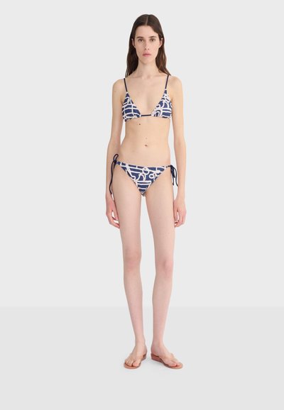 Femme debout, tournée vers l'avant, portant un bikini à motif bleu marine et blanc avec des liens sur les côtés, des bretelles fines et des sandales plates transparentes.