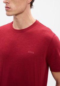 Červené bavlnené tričko s krátkymi rukávmi, okrúhlym výstrihom a jemným vyšitým logom "BOSS" na hrudi. Mäkká textúra a hladký povrch látky.