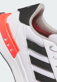 White athletic shoe with black stripes, gray accents, and a bright orange heel clip. Features a textured fabric upper and white sole.