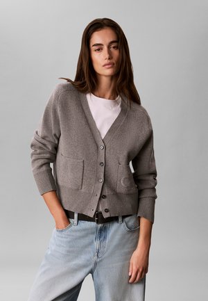 Femme aux longs cheveux foncés portant un cardigan gris boutonné par-dessus une chemise blanche et un jean bleu clair, debout avec une main dans la poche.