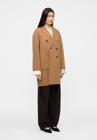 Δίπλο πέτο καμηλό παλτό από απαλό ύφασμα, με μαύρα κουμπιά, μεγάλες τσέπες και χαλαρή σιλουέτα. Συνδυάζεται με πλατύχωρα σκούρα παντελόνια.